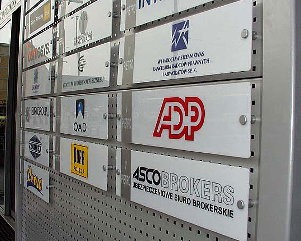 Tablica z lakierowanej aluminiowej blachy aluminiowejj ustawionej 
przed biurowcem ASCO z szyldami firm